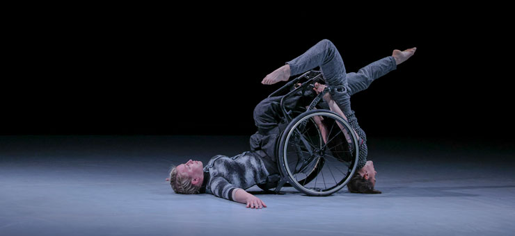 Två kvinnliga dansare. Den ena är rullstolsburen och ligger på golvet, sittandes i rullstolen. Den andra står på huvudet, med ryggen mot rullstolen , håller fast i den och pekar med benen framåt och bakåt.