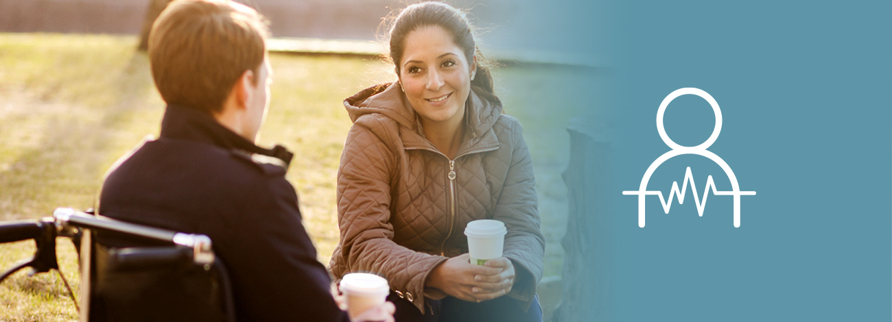 En kvinna sitter utomhus på en bänk och ler mot en annan person som hon dricker kaffe med