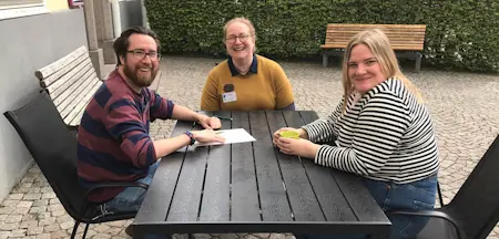 Martin Bensch från Helsingborgs stadsbibliotek, Linda Harrisson från Kävlinge bibliotek och Elin Halin från Lunds stadsbibliotek sitter runt ett mörkt trädgårdsbord. Alla tre skrattar och tittar in i kameran.