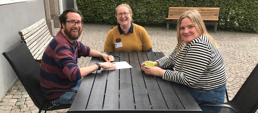 Martin Bensch från Helsingborgs stadsbibliotek, Linda Harrisson från Kävlinge bibliotek och Elin Halin från Lunds stadsbibliotek sitter runt ett mörkt trädgårdsbord. Alla tre skrattar och tittar in i kameran.
