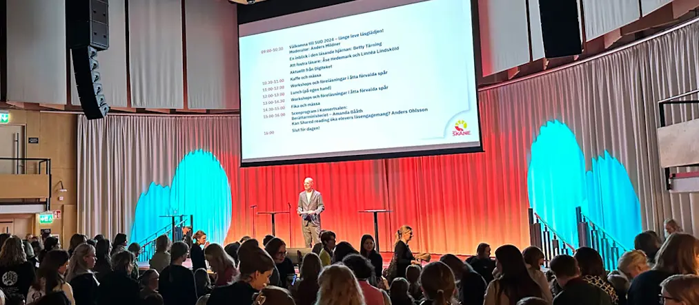 Moderatorn Anders Mildner syns på håll stå på en upplyst scen mitt emellan två ståbord. Ovanför honom syns en stor projektorduk med oläslig text från ett bildspel. Framför syns siluetter från en stor publik.