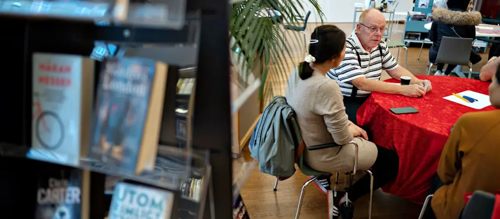 En äldre man i randig tröja och hängslen sitter och samtalar med en kvinna i beige kläder som man bara ser ryggen på. De sitter kring ett runt bord med röd duk. Fler människor runt bordet skymtas. I framgrunden syns en oskarp bokhylla med böcker.
