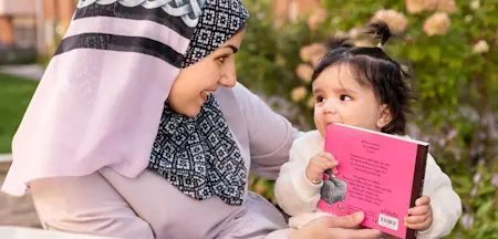 En kvinna i en rosa huvudduk med svart-vitt mönster ler och har ögonkontakt med ett litet barn med en liten tofs mitt uppe på huvudet. Barnet håller i en starkt rosa bok samtidigt som hen har ena bokhörnet i munnen.