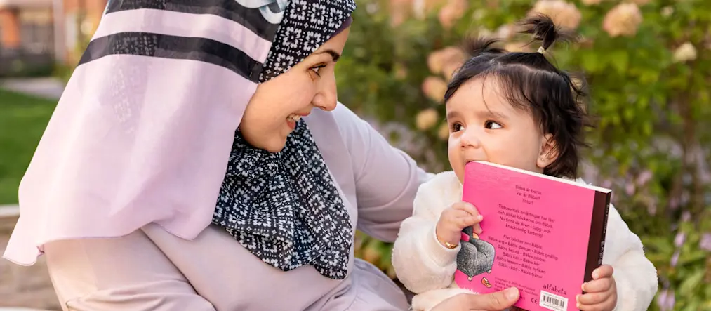 En kvinna i en rosa huvudduk med svart-vitt mönster ler och har ögonkontakt med ett litet barn med en liten tofs mitt uppe på huvudet. Barnet håller i en starkt rosa bok samtidigt som hen har ena bokhörnet i munnen.