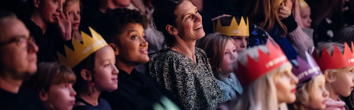 Vuxna och barn sitter tillsammans som en publik och tittar på en teaterföreställning på Malmö Stadsteater. Några av barnen har pappkronor på huvudet. De ser glada ut.