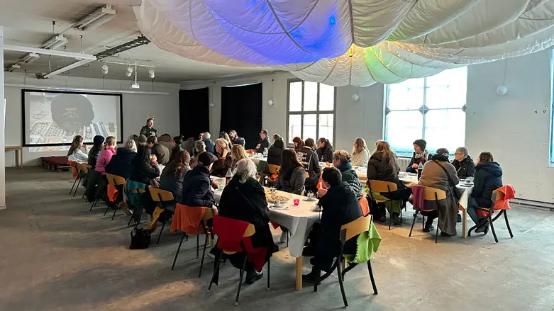 Runt två långbord i ett stort och avskalat rum med vita väggar och grått golv sitter ca 30 personer vända mot en stående person och bakom hen en stor bildduk. På ena väggen sitter stora fönster på rad. I taket hänger en gigantisk textillampa som lyser i rött, blått, gult och grönt.