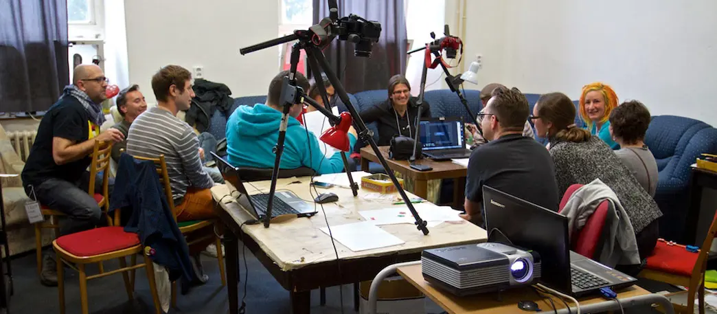 En grupp vuxna människor sitter i och runt en stor blå hörnsoffa. På ett soffbord i mitten och ett större bord bakom dem står två stativ med filmkameror. I förgrunden står ett annat bord med en uppslagen laptop och en lysande bildprojektor.