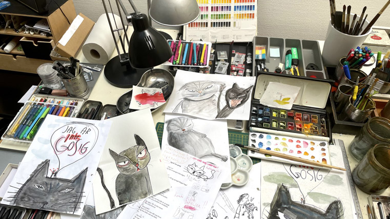 Skisser av katter på ett skrivbord med penslar, färg och pennor.
