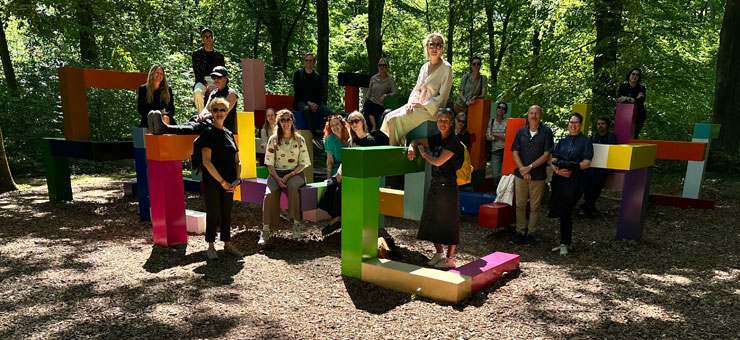 I en skogsglänta står en skulptur i olika färger. Skulpturen består av block som är staplade på varandra på olika sätt. Vid och på blocken står en grupp människor. 