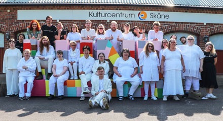 27 människor sitter och står på och bakom en skulptur i form av bokstäverna LUND. De flest har vita kläder. Alla ser glada ut. På väggen i bakgrunden står texten "Kulturcentrum Skåne". 