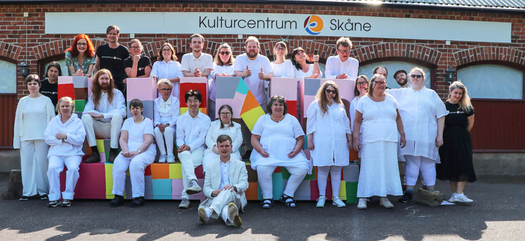 27 människor sitter och står på och bakom en skulptur i form av bokstäverna LUND. De flest har vita kläder. Alla ser glada ut. På väggen i bakgrunden står texten "Kulturcentrum Skåne". 