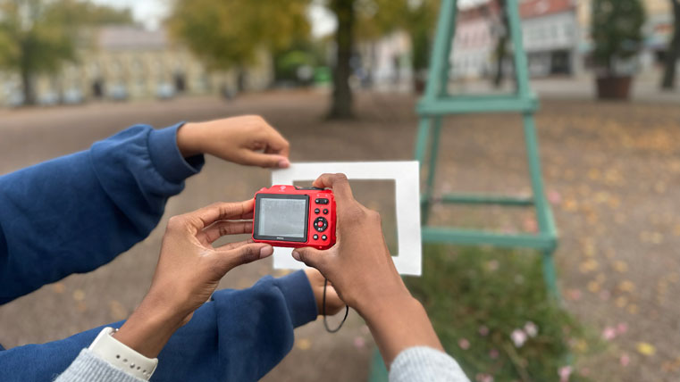 Två personer fotograferar utomhus med en liten röd digitalkamera. De håller upp en vit ram framför motivet på ett torg.