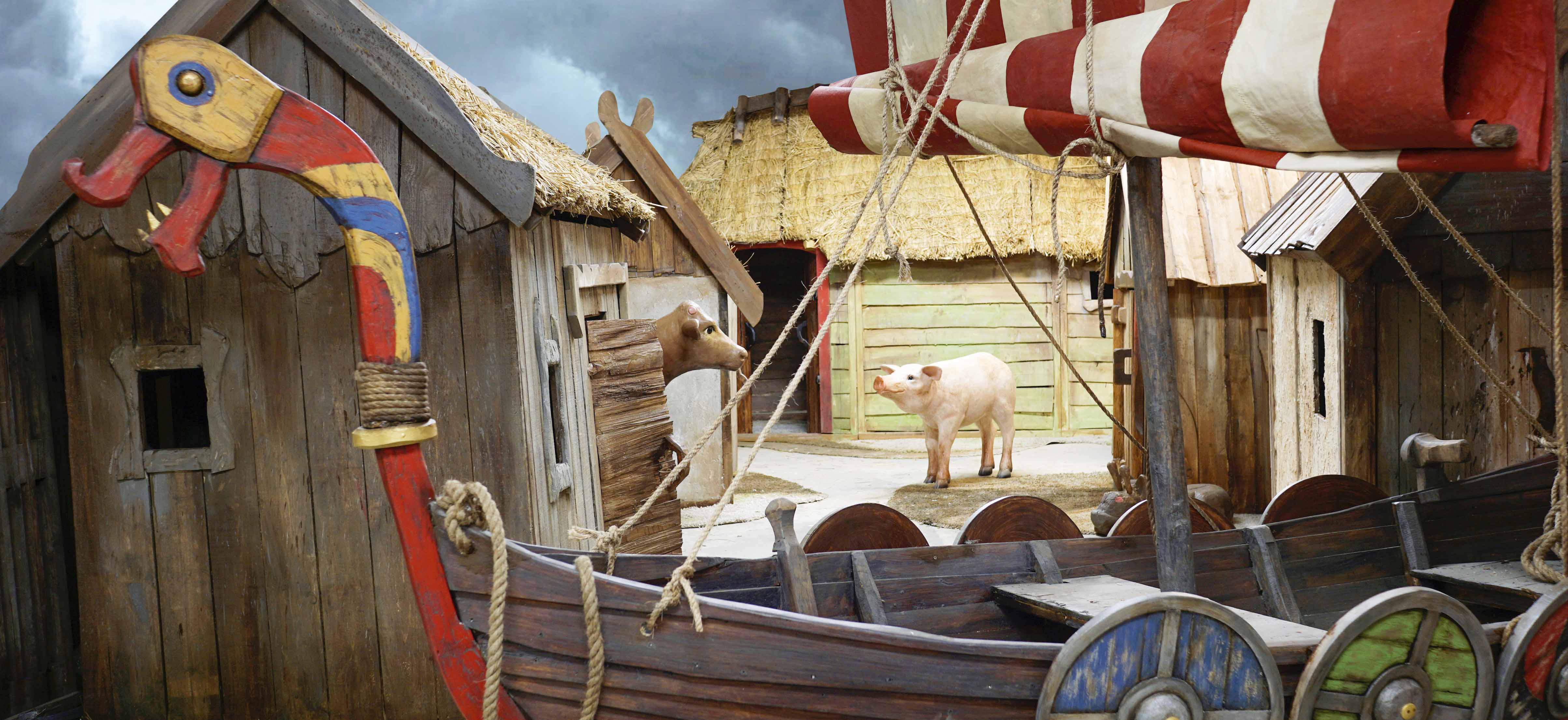 Bild på en uppbyggd utställning av en vikingaby. På bilden syns små hus med halmtak, en båt med sköldar på sidan i olika färger. En gris och en ko i mitten.