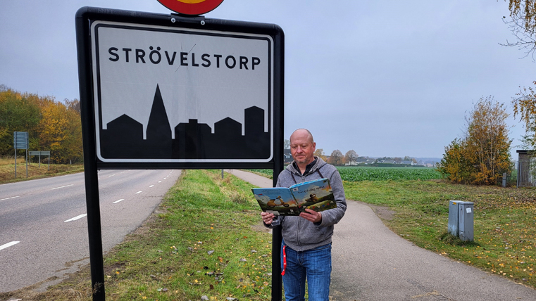 Man som läser en bok bredvid en vägskylt med texten Strövelstorp.