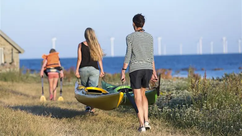 Två personer bär på en båt vid havet och i bakgrunden syns vindkraftverk.