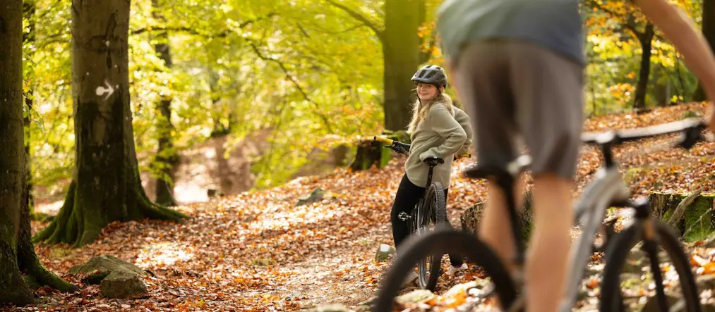 Flicka står lutad mot cykel i bokskog och tittar på cyklande kompis.