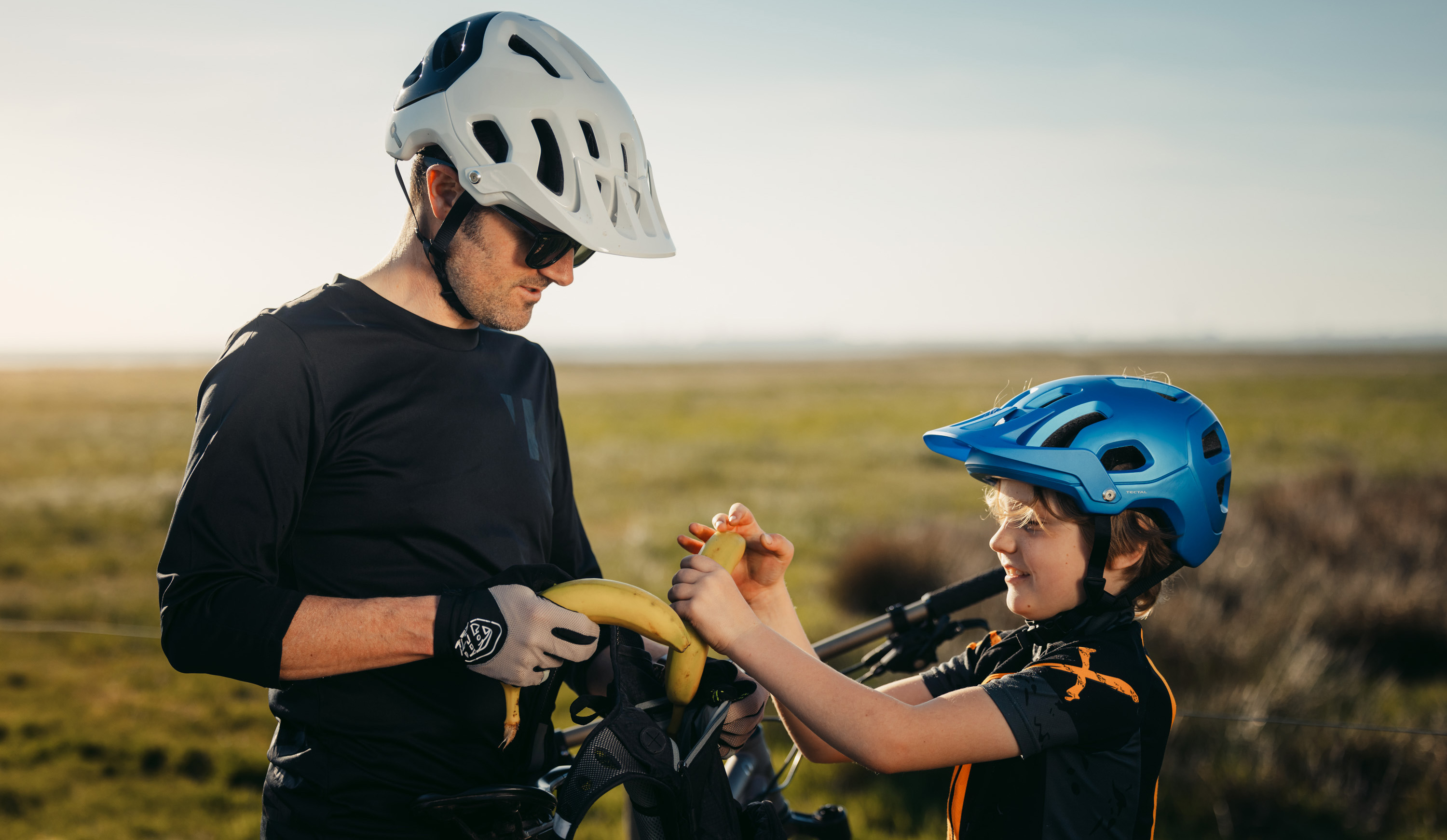 En vuxen man med cykelhjälm på huvudet bjuder ett barn med hjälm på huvudet på banan. 